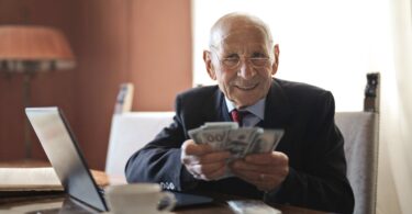 confident senior businessman holding money in hands while sitting at table near laptop